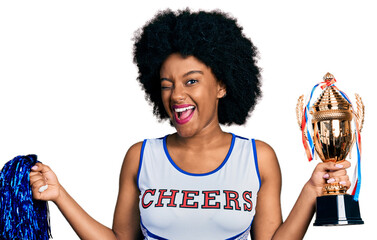 Young african american woman wearing cheerleader uniform holding pompom and trophy winking looking...