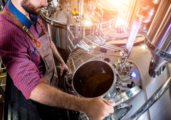 Young male brewer in leather apron supervising the process of beer fermentation at modern brewery factory