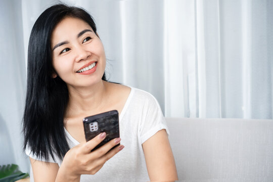 smiling Asian woman using a smartphone, thinking and daydreaming of something with happy face