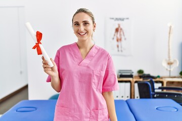 Young blonde woman wearing physiotherapist uniform holding diploma at physiotherapy clinic
