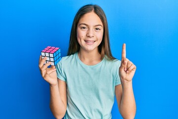 Beautiful brunette little girl playing colorful puzzle cube intelligence game smiling with an idea or question pointing finger with happy face, number one