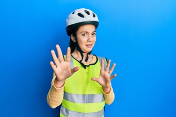 Beautiful brunette little girl wearing bike helmet and reflective vest afraid and terrified with fear expression stop gesture with hands, shouting in shock. panic concept.
