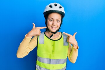 Beautiful brunette little girl wearing bike helmet and reflective vest smiling cheerful showing and pointing with fingers teeth and mouth. dental health concept.