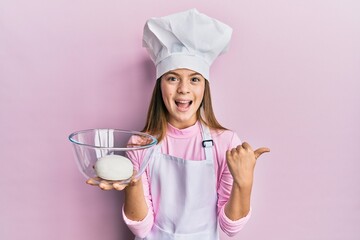Beautiful brunette little girl wearing chef hat holding bread dough pointing thumb up to the side...