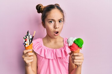 Beautiful brunette little girl eating ice cream cones in shock face, looking skeptical and...