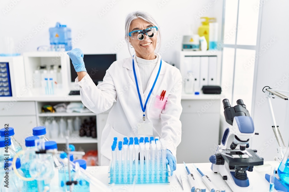 Poster middle age grey-haired woman wearing scientist uniform pointing thumb up to the side smiling happy w