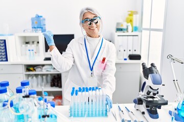 Middle age grey-haired woman wearing scientist uniform pointing thumb up to the side smiling happy with open mouth
