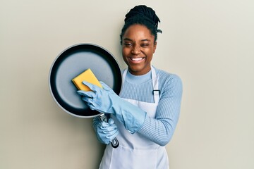 African american woman with braided hair wearing apron holding scourer washing pan winking looking...
