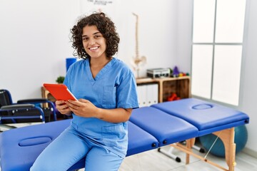 Young hispanic woman wearing physio therapist uniform using touchpad at clinic