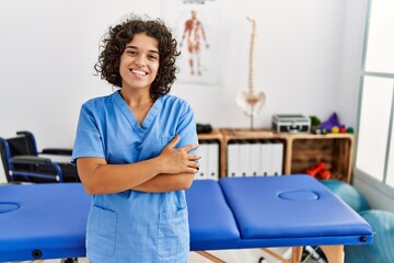 Young hispanic physiotherapist woman working at pain recovery clinic happy face smiling with crossed arms looking at the camera. positive person. - obrazy, fototapety, plakaty
