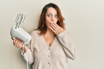 Young caucasian woman holding food processor mixer machine covering mouth with hand, shocked and...