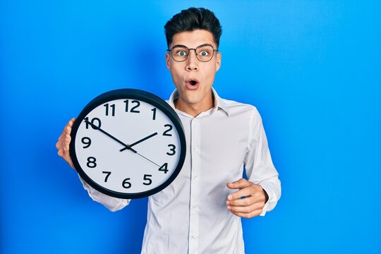 Young hispanic man holding big clock scared and amazed with open mouth for surprise, disbelief face