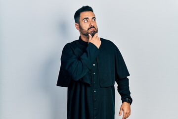 Handsome hispanic man with beard wearing catholic priest robe thinking worried about a question,...