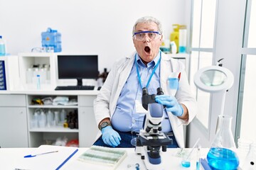 Senior caucasian man working at scientist laboratory in shock face, looking skeptical and sarcastic, surprised with open mouth
