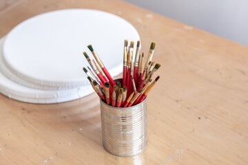 Red paint brushes in a metal can on a table with round canvases for painting.