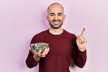 Young bald man holding sunflower seeds bowl smiling happy pointing with hand and finger to the side