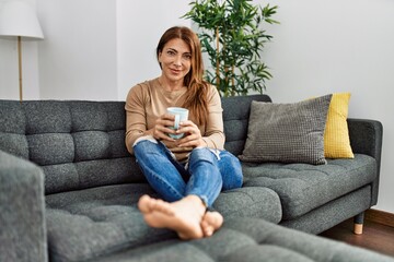 Middle age caucasian woman drinking coffee sitting on the sofa at home.