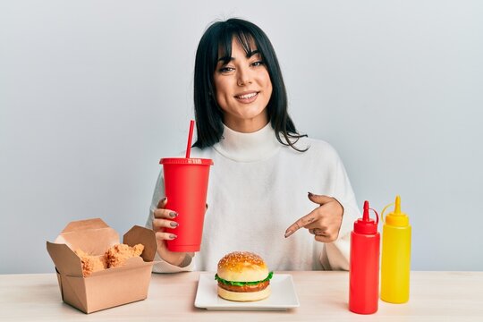 Young brunette woman with bangs eating a tasty classic burger with ketchup and mustard smiling happy pointing with hand and finger