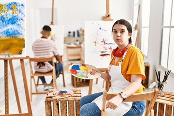 Young artist woman painting on canvas at art studio puffing cheeks with funny face. mouth inflated...