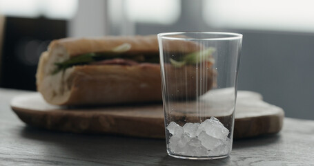 pour crushed ice in tumbler glass on oak table with sandwich on background