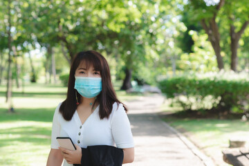 Fototapeta premium attractive business woman wearing face mask hold smartphone standing in park with sunrise and blur nature background. business new normal with corona virus or covid-19.