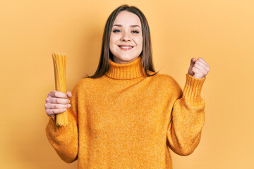 Young hispanic girl holding uncooked spaghetti screaming proud, celebrating victory and success very excited with raised arm