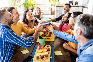 Smiling multiracial group of young people having breakfast, laughing and cheering with hot drinks together at coffee bar - Diverse millennial colleagues enjoying meal during work break - Focus on cups