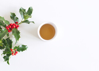 Christmas styled composition, decorative frame, banner. Holly tree green leaves, red berries, branches and white porcelain mug with tea on white table background. Flat lay, top view.