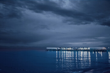 Fototapeta na wymiar Twilight landscape of pier stretching out into sea. Cloudy sky. Illumination and umbrellas on ocean bridge. Nature summer landscape. Tourism background. Travel and vacation. Glare from lamps on water.