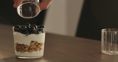 add blueberries to granola with yogurt in tumbler glass on walnut table