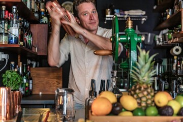 View of a professional bartender shaking a cocktail behind the counter of a tiny small bar wearing a casual shirt, young businessman concept.