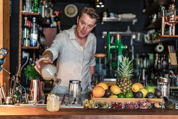 Fototapeta premium View of a professional bartender pouring a cocktail from a glass jar into a glass with lemon and ice, view of a small cocktail bar with fruits on counter, Lisbon, Portugal.