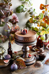 Chocolate cheesecake with fresh figs on a high wooden stand among autumn raspberry leaves. Still life with sweets and natural materials.