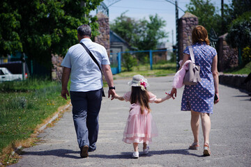 Obraz premium family walking in the park. mom dad and little girl walking holding hands. back view. daughter in a pink dress. friendly loving family concept, love. outdoor, walk in the fresh air, threesome