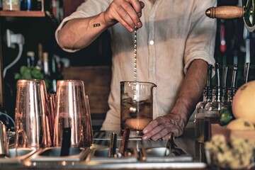 An expert bartender mixing a cocktail in a glass mixer with a long spoon, standing behind the counter of his beautiful cocktail bar in a night club.