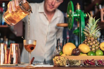 A professional bartender pouring a cocktail in a glass from a shaker and mixer, view of a barman working in a bar.