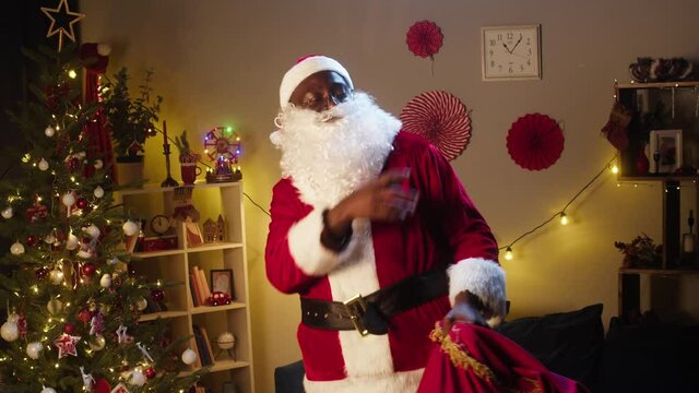 Santa Claus dancing in living room, holding big bag full of gifts. African american man in costume of Santa having fun, showing dance. New year time and christmas holidays concept. 