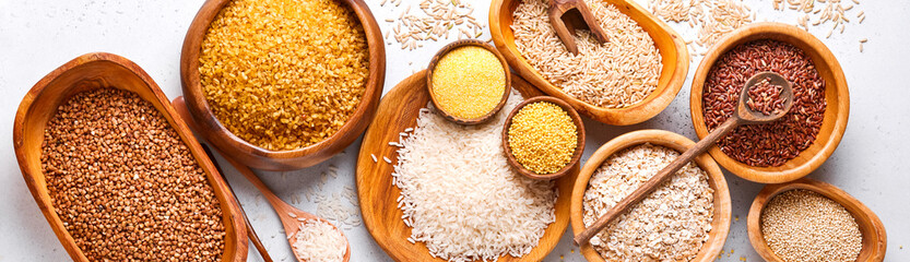 White, brown and red rice, buckwheat, millet, corn groats, quinoa and bulgur in wooden bowls on the light gray kitchen table. Gluten-free cereals. Top view with copyspace. Banner.