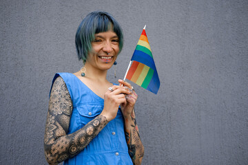 Beautiful smiling young woman with shirt blue hair, wearing blue dress holding little rainbow flag...