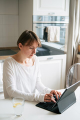 Young lady with short hair wearing open shoulder t shirt working on laptop sitting at kitchen table.