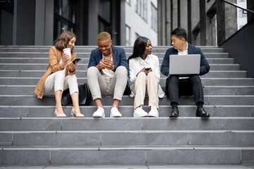 Fototapeta premium Multiracial business team talking and using digital devices on stairs in city. Concept of business cooperation. Idea of remote and freelance work. Modern successful business people in formal wear