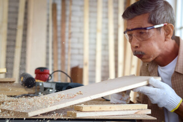 Elderly carpenter wearing safety glasses goggles blowing sawdust off piece of wood plank, senior...