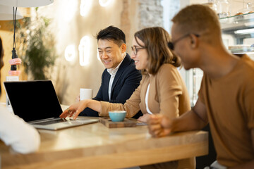 Multiracial business team watching something on laptop while working in cafe or restaurant. Concept of remote and freelance work. Idea of business cooperation. Smiling people sitting at wooden table