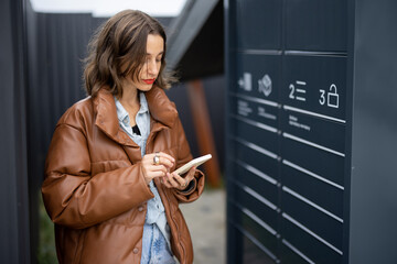 Young caucasian woman using smartphone near automatic post terminal. Focused girl on city street....