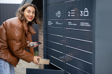 Young caucasian woman putting parcel to cell of automatic post terminal. Smiling girl holding...