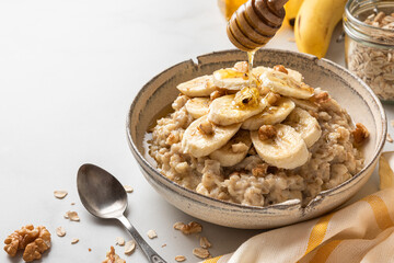 Pouring honey in a bowl of oatmeal porridge with banana and walnuts. Healthy diet breakfast