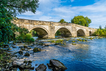 A view along the rocky shore towards an old medieval bridge over the River Ure on the outskirts of Ripon, Yorkshire, UK in summertime