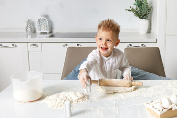 The boy at the table in the white kitchen cheerfully prepares a pie from dough, eggs, milk and flour