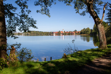 Obraz premium View over an idyllic lake in Bavaria with reflective village including white church tower. In the foreground trees with shadows at a bathing area