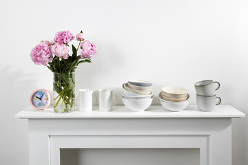 Bouquet of pink peonies, a stack of cups, plates, bowls, white chest of drawers in the interior of the kitchen